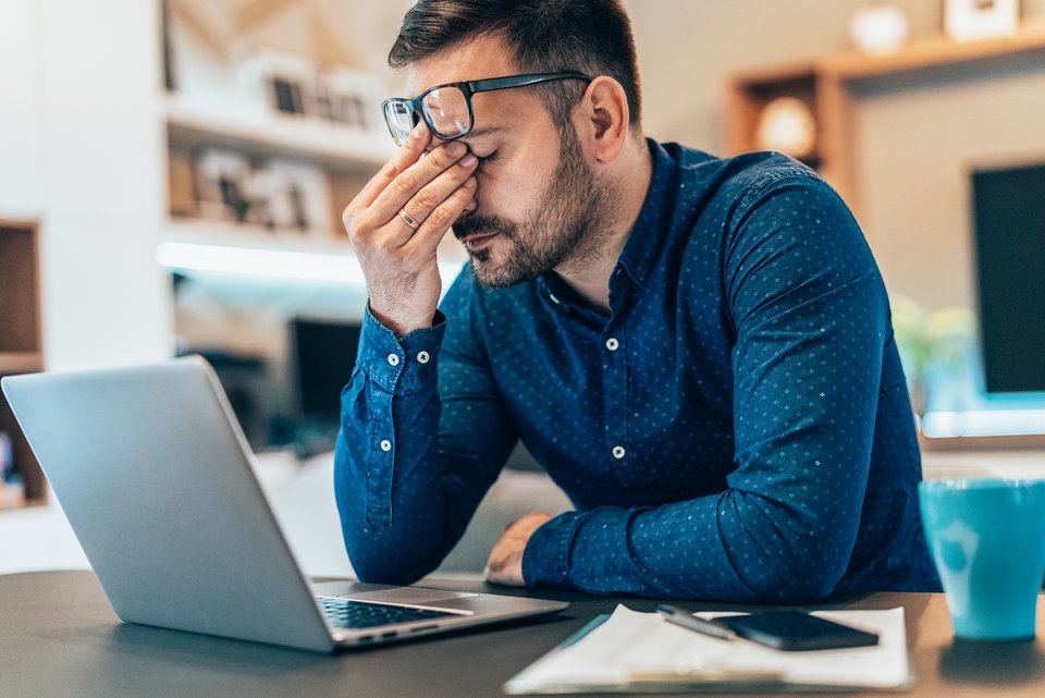 Ein Mann, der Freiberufler ist, sitzt vor seinem Laptop, schiebt die Brille auf die Stirn und reibt sich müde die Augen, weil er keine Ideen mehr hat