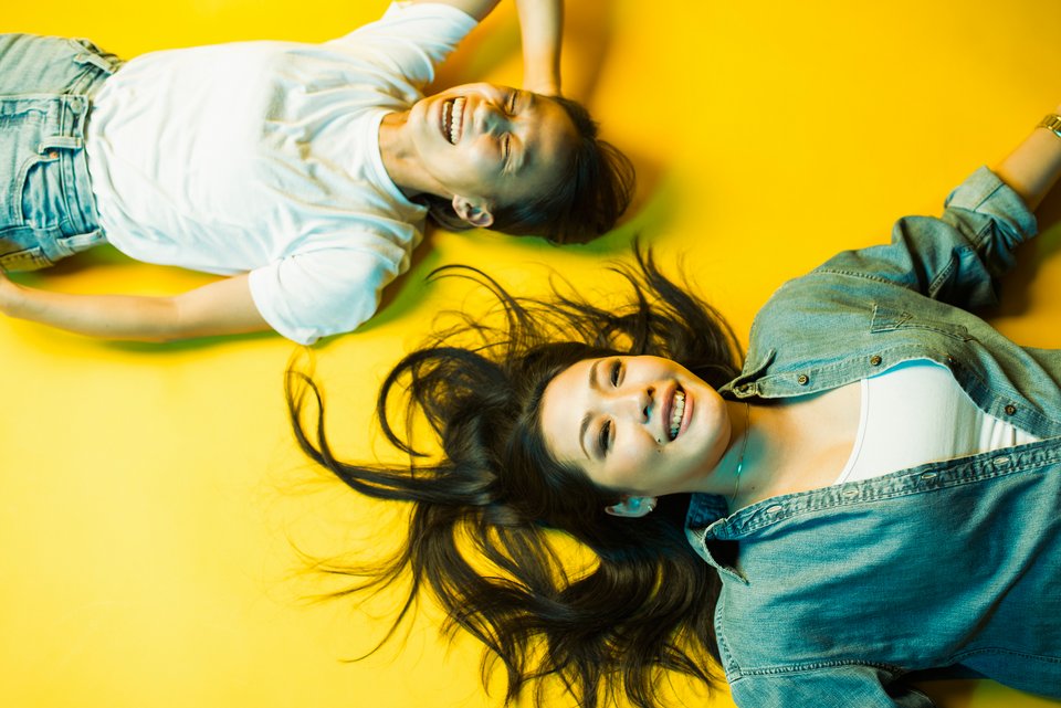 Zwei junge Frauen aus Japan liegen auf einem gelben Boden und lachen ausgelassen