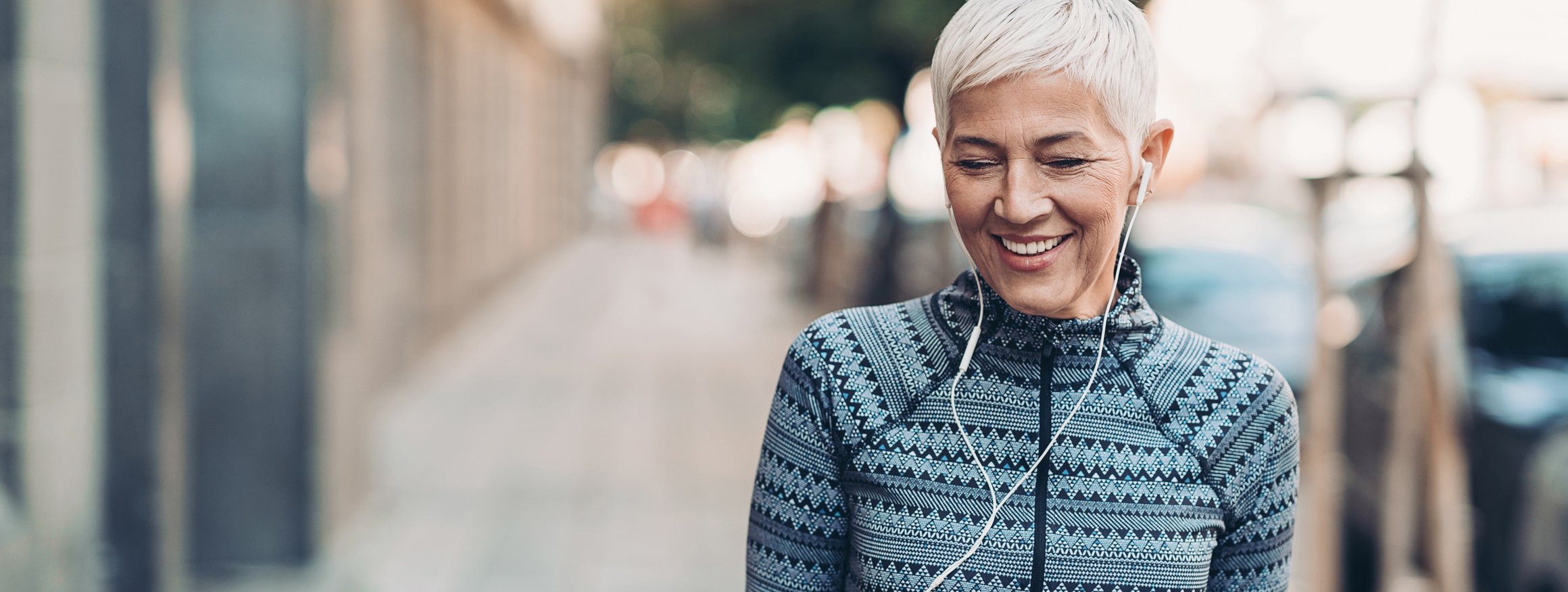Eine ältere Frau mit grauen kurzen Haaren, läuft in Sportkleidung  und In-Ears lächelnd durch eine Straße