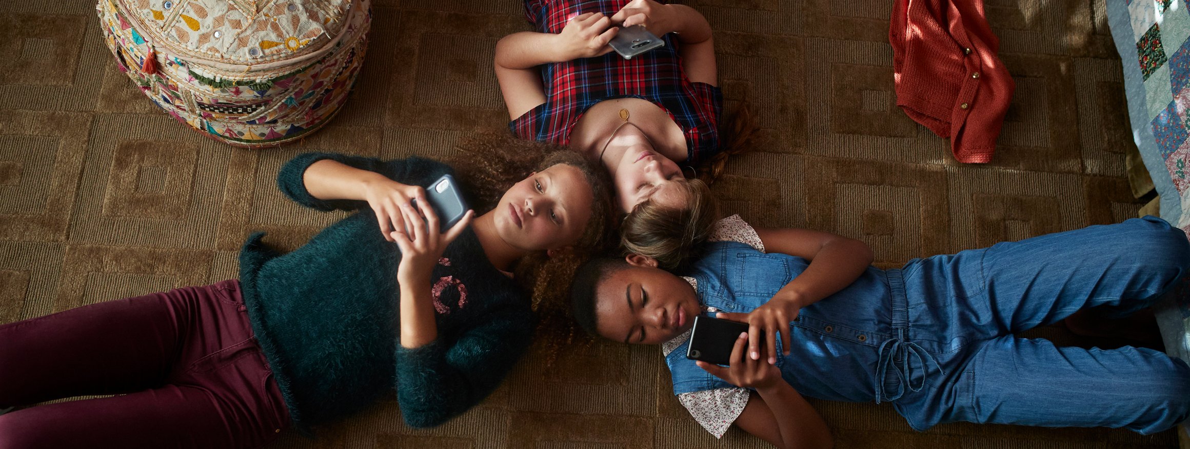 Das Foto zeigt, drei Kinder, die auf dem Boden eines Kinderzimmers liegen. Alle halten ihre Smartphones in den Händen und tippen darauf.