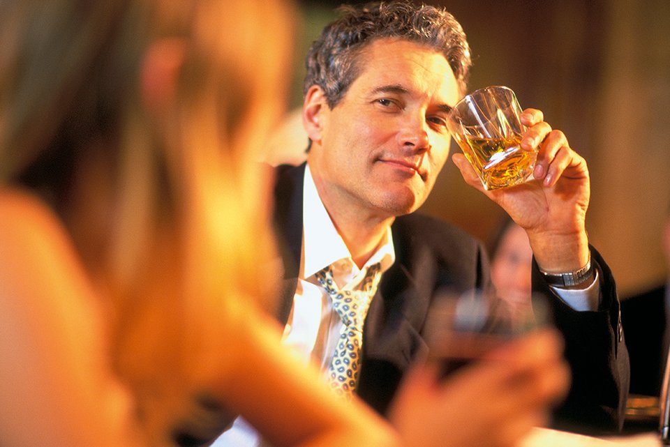 Ein Mann im Anzug hält an einem Thresen in einer Bar lässig einen Drink in der Hand und schaut überlegen zu einer Frau hin