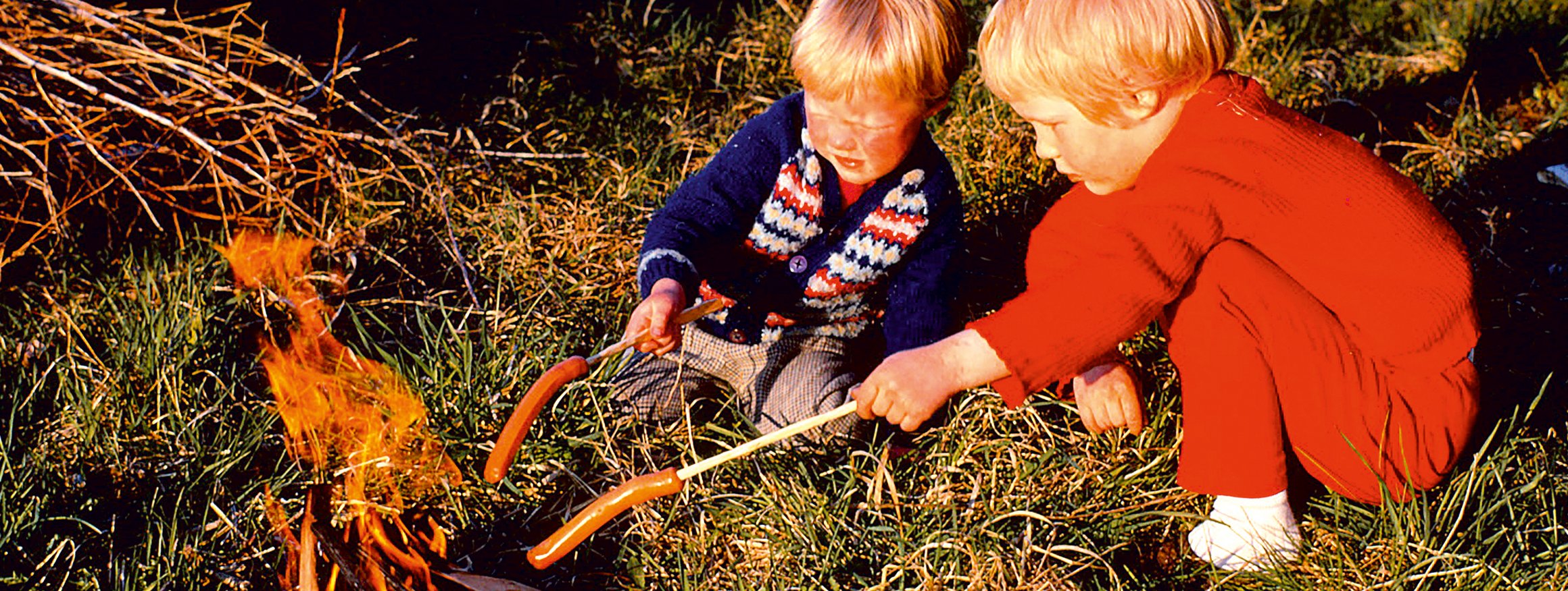Ein älteres Foto zeigt zwei blonde Kinder auf einer Wiese mit Würstchen auf Stöcken gespießt vor einem Lagerfeuer