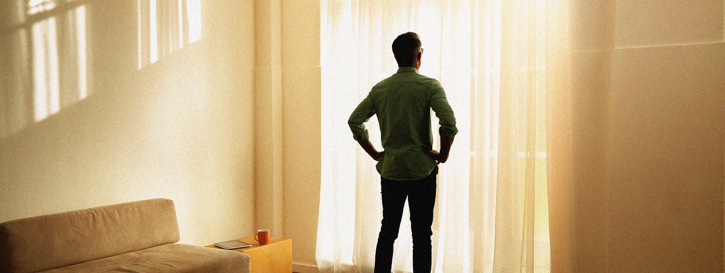 Ein älterer Mann steht traurig in einem Raum und schaut aus dem Fenster, die Hände an den Hüften