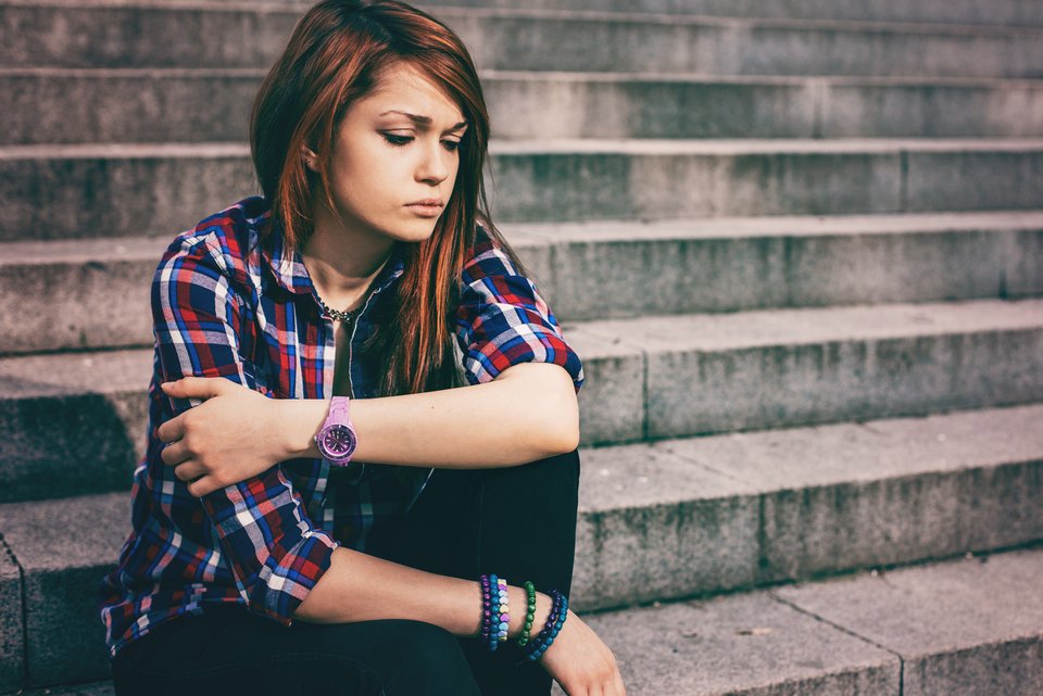 Eine Jugendliche mit langen Haaren und Karohemd sitzt allein und traurig auf den Stufen einer Betontreppe und ist depressiv
