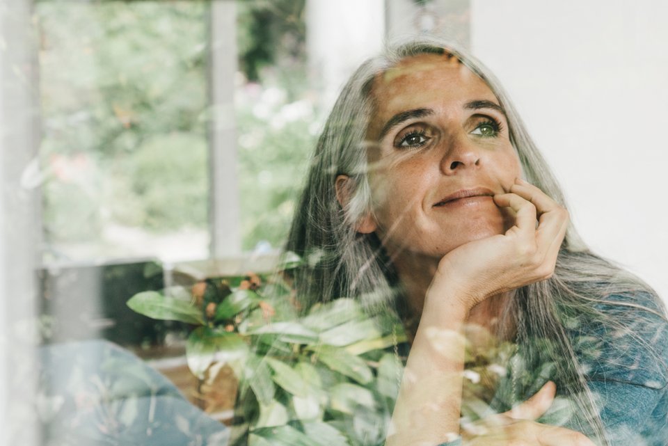 Eine ältere Frau mit langen grauen Haaren schaut sehnsüchtig auch dem Fenster und träumt von dem perfekten Leben