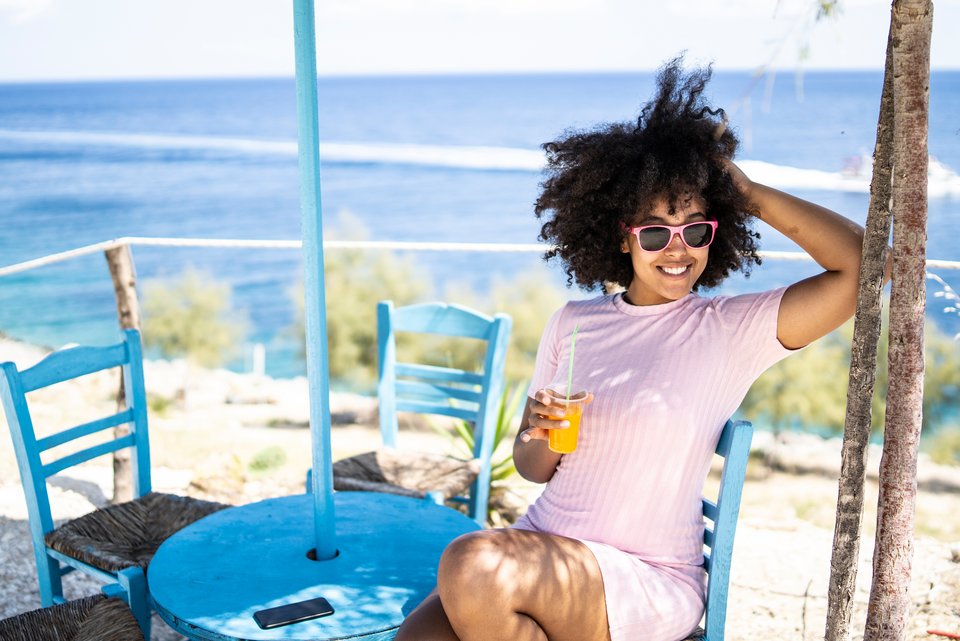 Eine junge Frau mit dunklem Lockenkopf und Sonnenbrille sitzt lachend mit einem Drink in der Hand an einer Strandbar