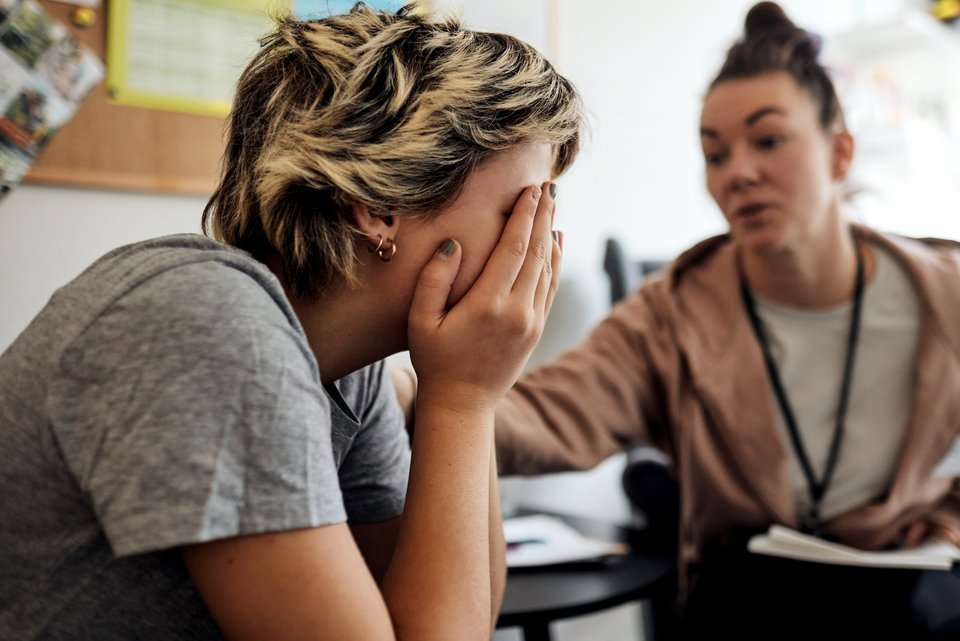 Eine Schülerin mit zweifarbiger Haarfrisur, Ohrring und lackierten Nägeln sitzt traurig vor der Psychologin und hält sich die Hände vor das Gesicht