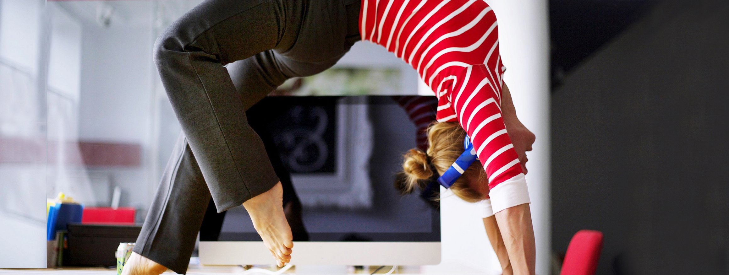 Eine junge Frau macht als Supertasker während ihrer Arbeit im Büro auf dem Schreibtisch vor ihrem PC akrobatische Yogaübungen