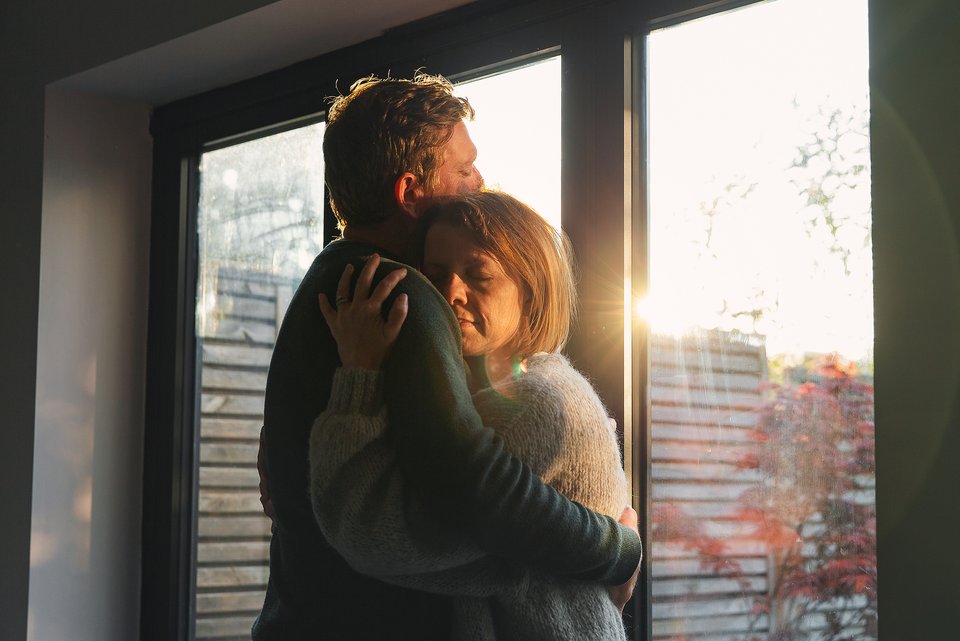 Mann und Frau umarmen sich und stehen vor einem Fenster durch das die Abendsonne scheint