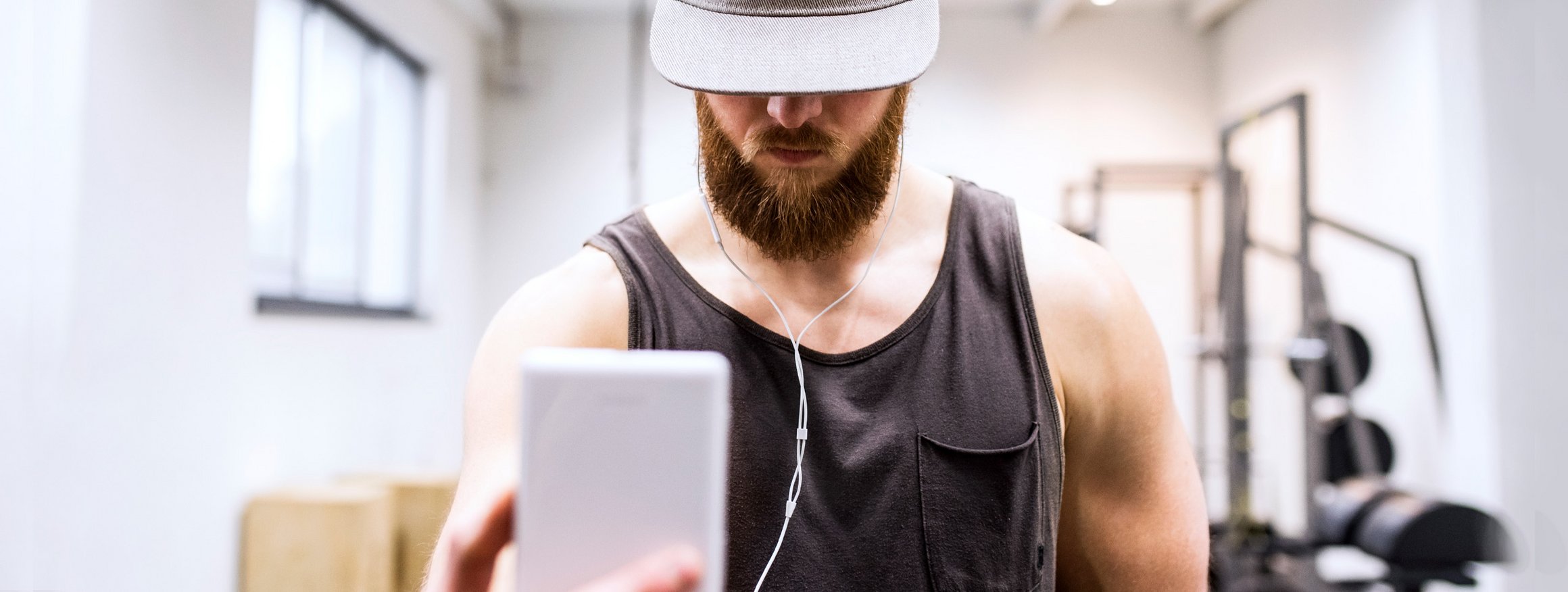 Ein muskulöser Mann im Fitnessstudio mit Baseballkappe und Bart macht mit seinem Smartphone ein Selfie 