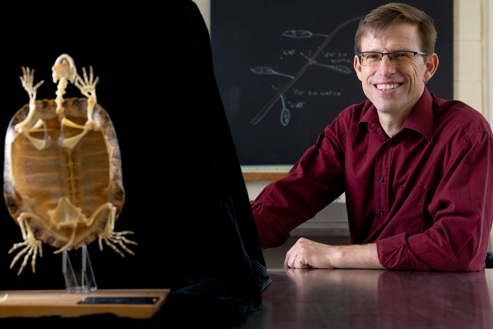 Der Forscher, Fritz Beithaupt, sitzt lächelnd vor einer Tafel , vor ihm steht ein präpariertes Fossil