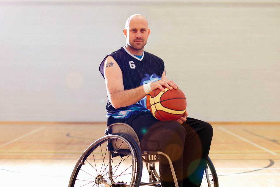 Ein junger, behinderter Sportler hat einen Basketball in der Hand und steht mit seinem Rollstuhl auf dem Spielfeld und meistert sein schweres Schicksal
