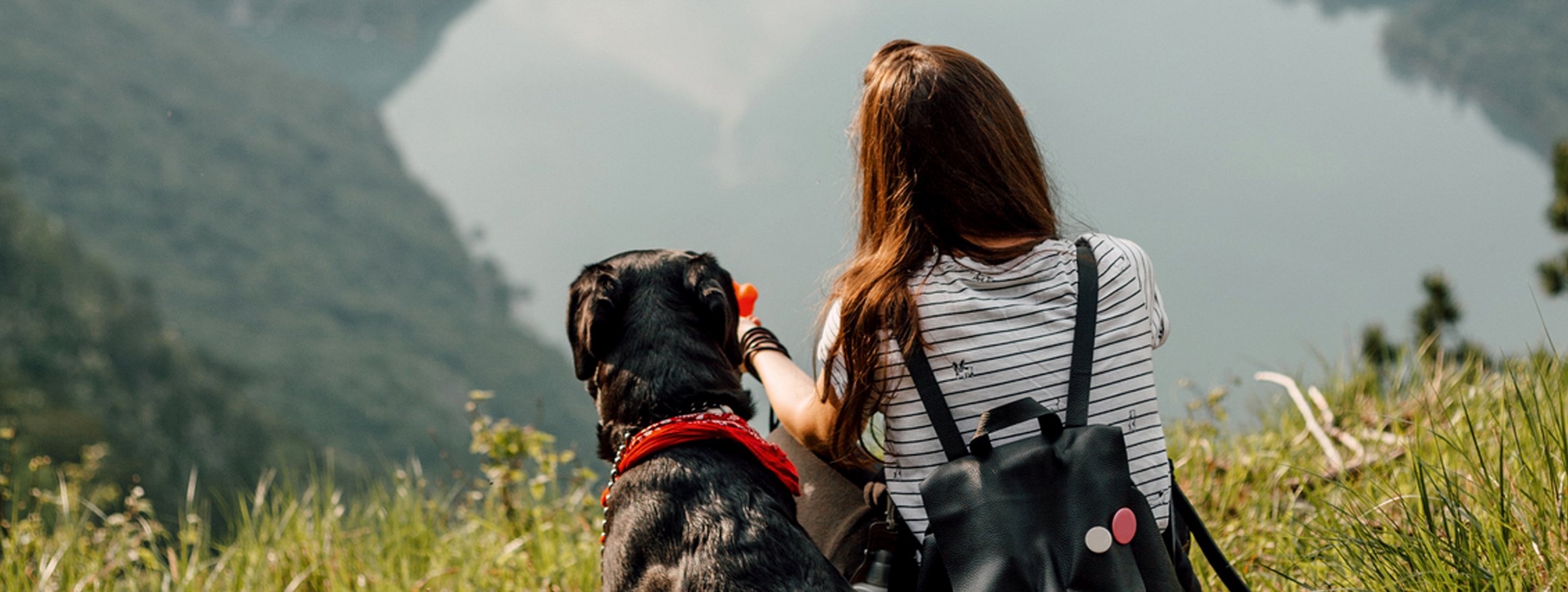 Eine Frau sitzt mit Rucksack auf einer Wiese zusammen mit ihrem Hund und genießt die schöne Aussicht in ein Tal mit einem See