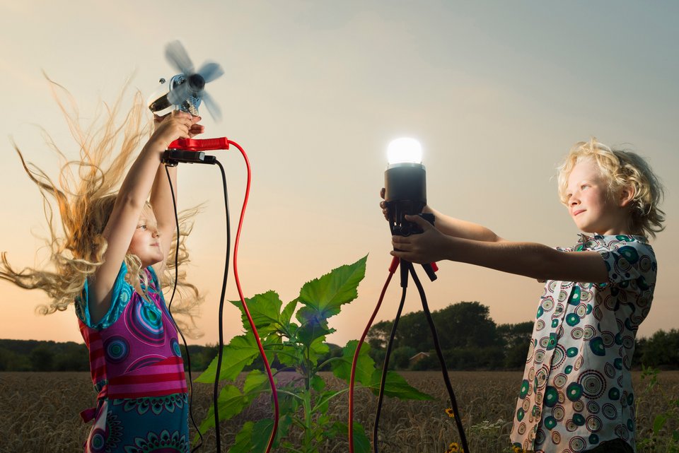 Zwei Kinder stehen an einem Feld mit Sonnenblumen und halten jeweils eine Lampe und einen Ventilator in den Händen, während sich bei dem Mädchen die Haare durch den Luftzug des Ventilators hochstellen