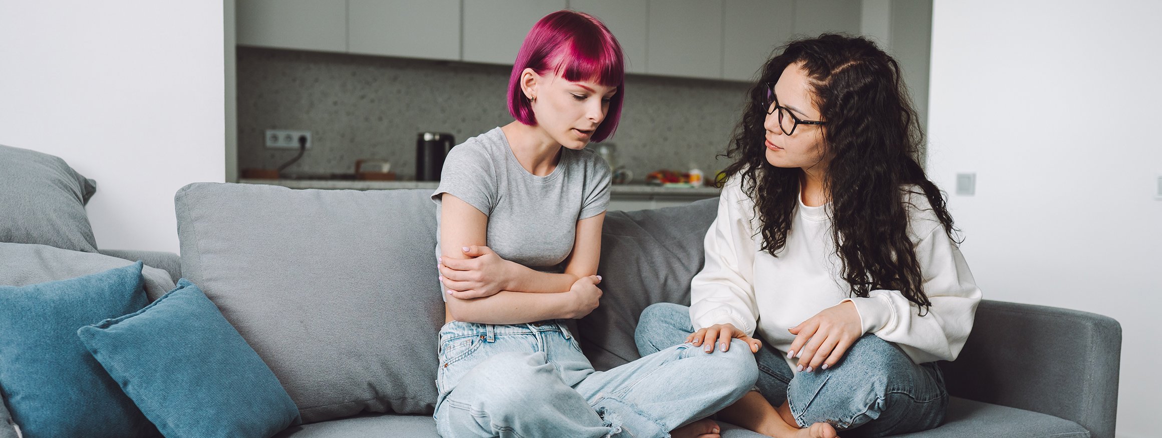 Eine Jugendliche mit pinken Haaren sitzt im Schneidersitz und verschränkten Armen auf der Couch. Ihre Mutter sitzt neben ihr und hört ihr zu.