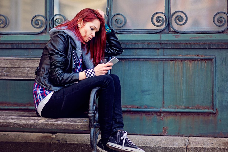 Eine Jugendliche mit roten langen Haaren, Lederjacke und Chucks, sitzt lässig auf einer Bank und schaut in sich gekehrt in ihr Smartphone