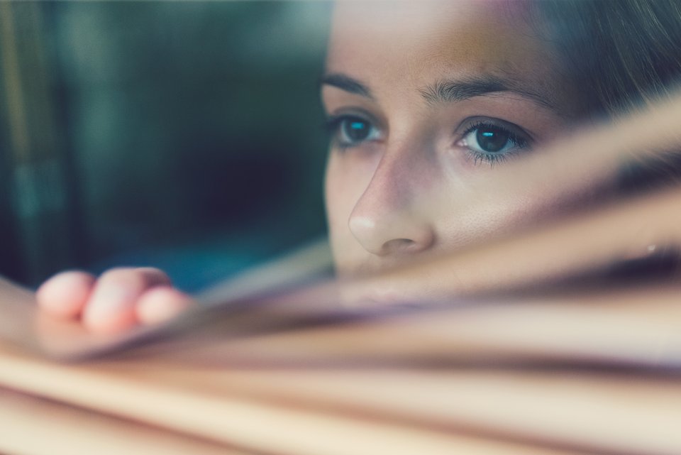 Eine Frau schaut unsicher an einer Jalousie aus dem Fenster und fürchtet sich vor Umwelteinflüssen, die sie für gefährlich hält