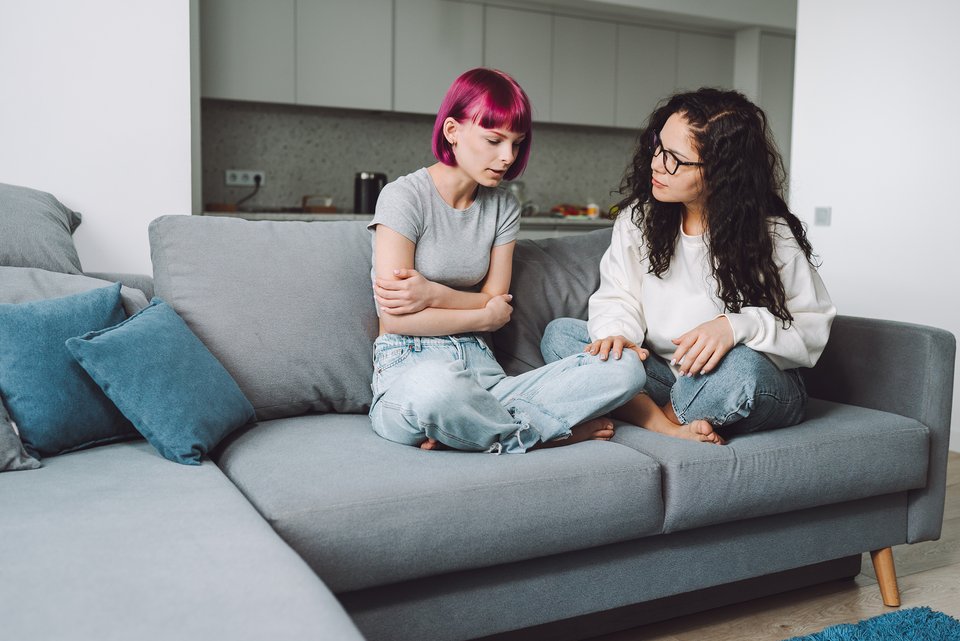 Eine Jugendliche mit pinken Haaren sitzt im Schneidersitz und verschränkten Armen auf der Couch. Ihre Mutter sitzt neben ihr und hört ihr zu.