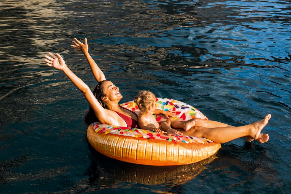 Eine Frau sitzt mit Badekleidung und ihrem Kind auf einer Schwimminsel im Wasser und reckt lachend die Arme nach oben