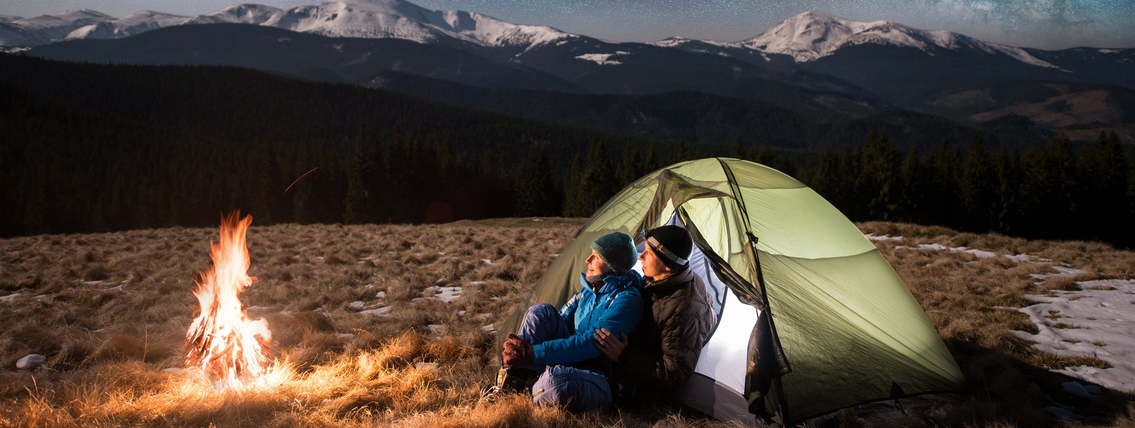Ein Paar sitzt vor seinem Zelt am Lagerfeuer und schaut dabei in den schönen Sternenhimmel vor einer Bergkulisse