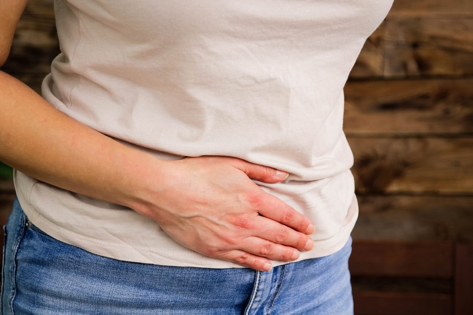 Eine Frau in Jeans und weißem T-Shirt hält sich mit der Hand den Bauch