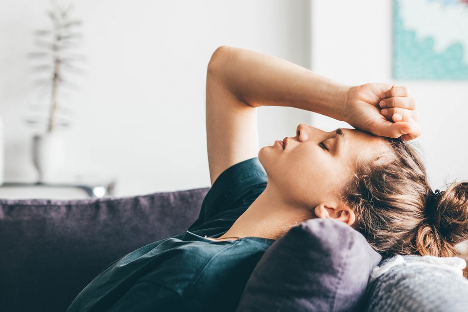 Eine junge Frau liegt auf der Couch, den Kopf auf ein Kissen gelegt, und hält sich leidend die Hand an die Stirn, während sie die Augen geschlossen hat