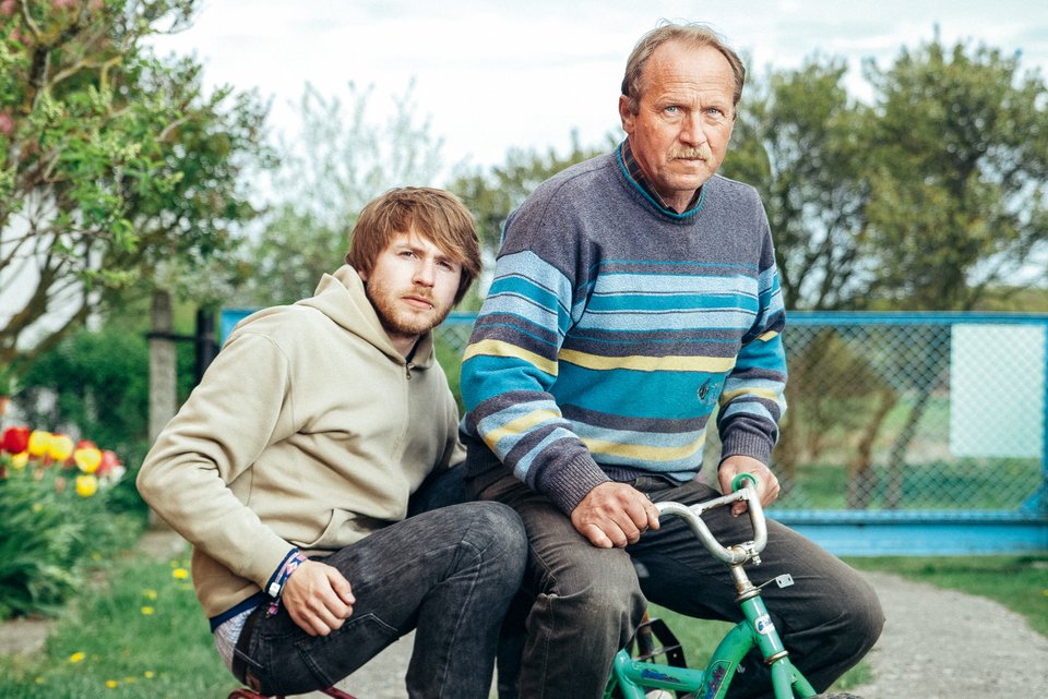 Ein Mann sitzt in der Hocke auf einem Dreirad, daneben ebenfalls auf einem Dreirad, sein erwachsener Sohn, der sich von seinen Eltern nicht ablösen kann