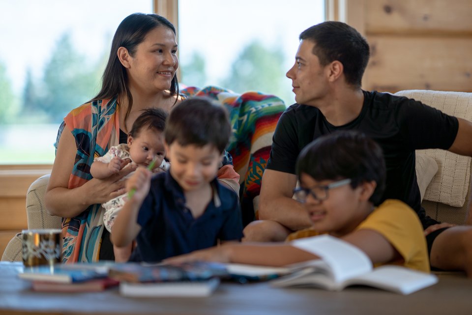 Eine Indigene Familie sitzt auf der Couch. Die Kinder spielen. Die Frau hat ein Baby auf dem Arm und lächelt ihren Mann verlegen an.