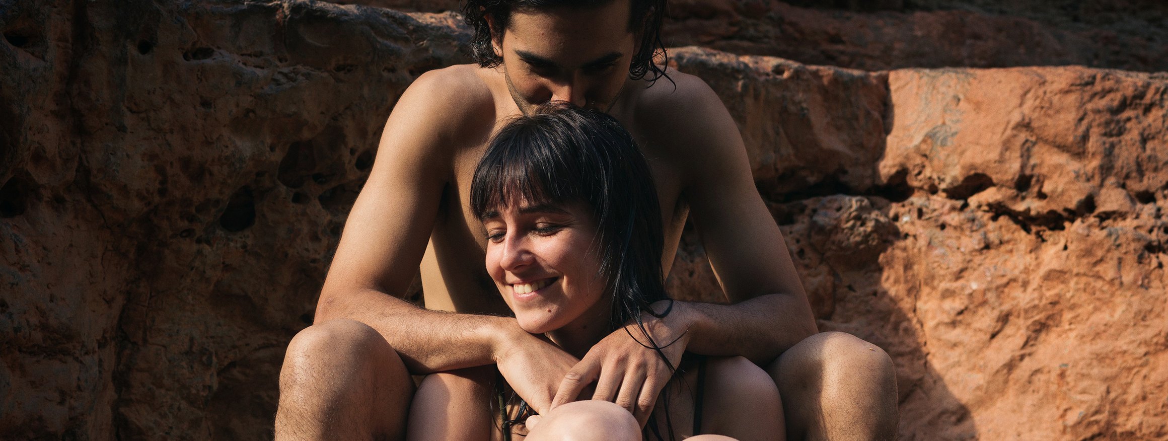 Ein junges nacktes Paar sitzt lachend vor einer Felswand, während er die Arme um ihre Schultern gelegt hat