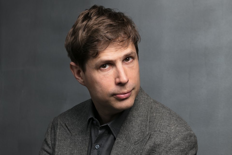 Der Schriftsteller Daniel Kehlmann sitzt vor grauem Hintergrund und schaut in die Kamera