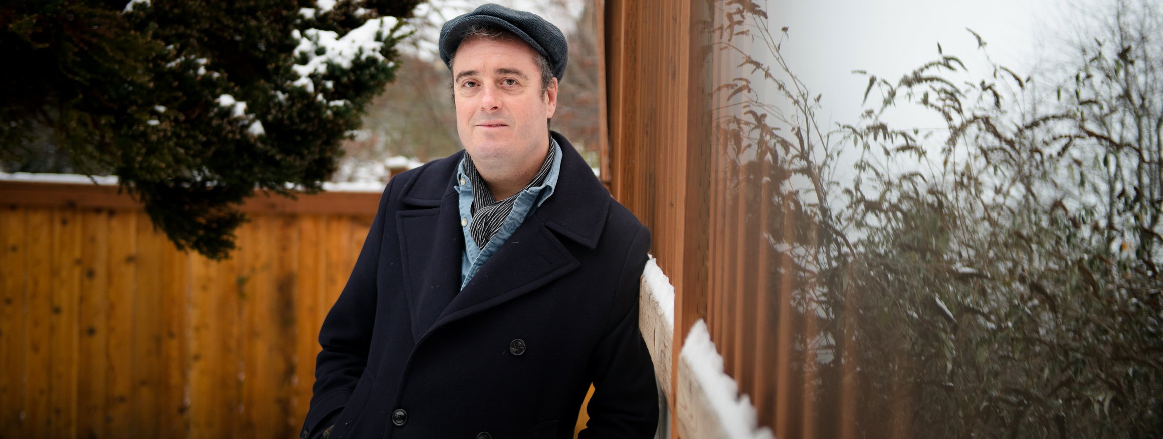 Der britische Psychologe Tim Lomas steht mit Mützen und Winterjacke auf einem Balkon, hinter ihm ein  schneebedeckter Garten mit Baum