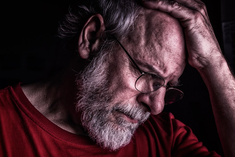 Ein älterer Mann mit Brille und rotem Shirt hält sich ernst eine Hand an den Kopf