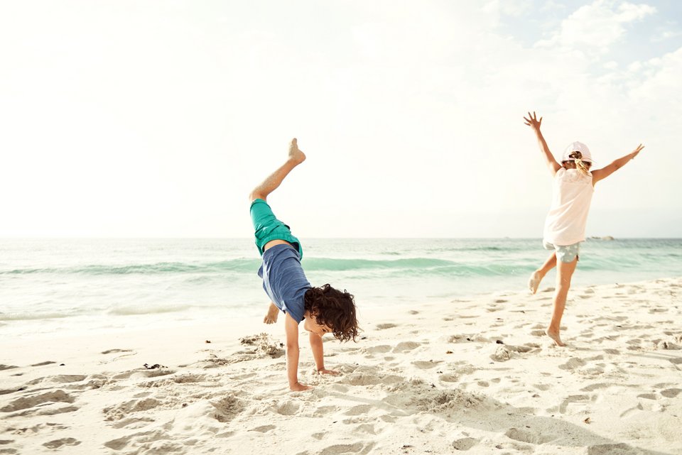 Zwei Kinder springen frei und ausgelassen an einem weißen Strand und der Junge schlägt dabei ein Rad