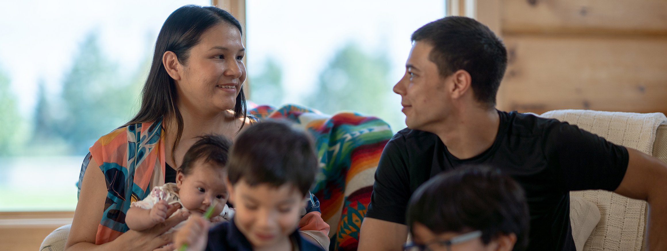 Eine Indigene Familie sitzt auf der Couch. Die Kinder spielen. Die Frau hat ein Baby auf dem Arm und lächelt ihren Mann verlegen an.