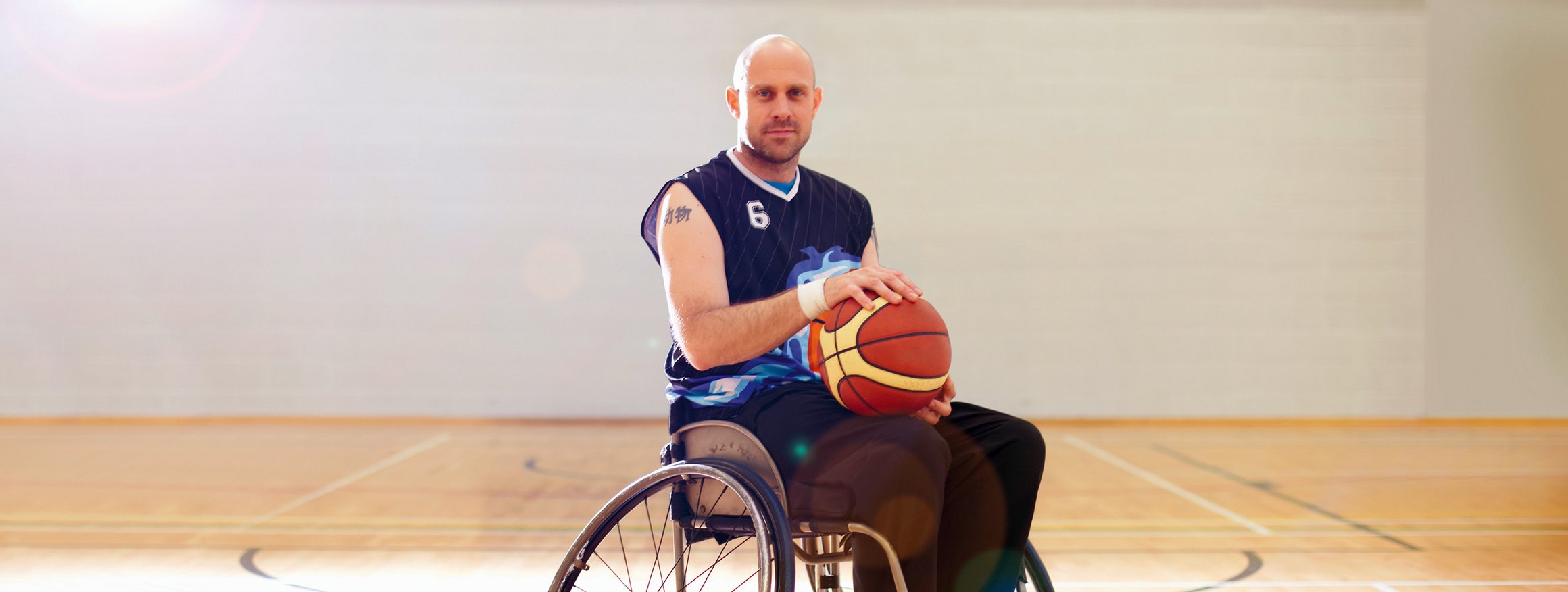 Ein junger, behinderter Sportler hat einen Basketball in der Hand und steht mit seinem Rollstuhl auf dem Spielfeld und meistert sein schweres Schicksal