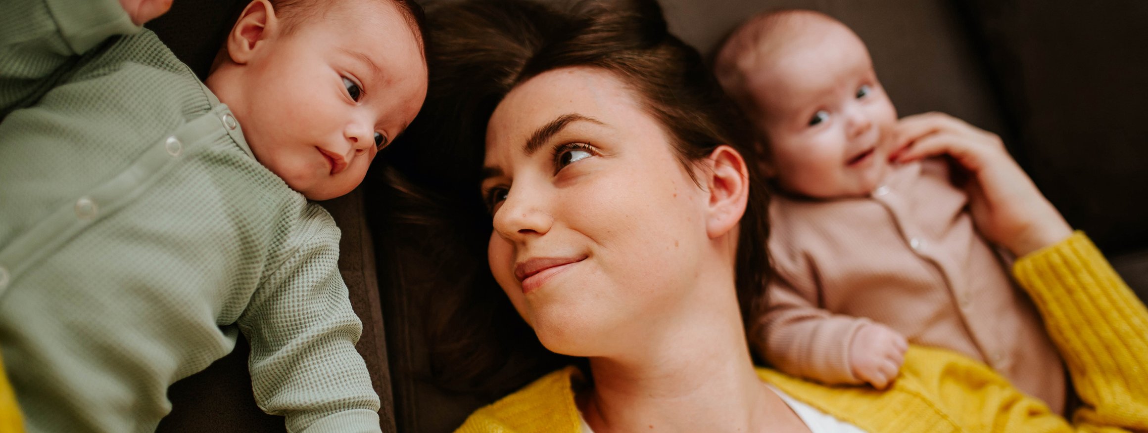 Eíne junge Mutter liegt zusammen mit ihren beiden Babies da und berührt sie sanft und schaut dabei liebevoll