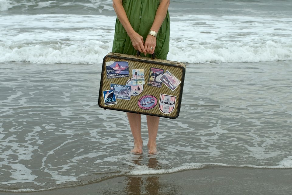 Eine Frau steht im grünen Kleid am Meer mit den Füßen im Wasser und hält dabei einen alten Reisekoffer in den Händen, der schon viele Aufkleber hat