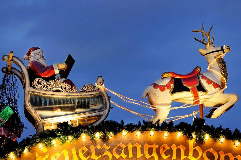 Auf einem Weihnachtsmarkt steht ein beleuchteter Stand mit einem Weihnachtsmann mit Geschenken in einem Schlitten mit Rentier auf dem Dach des Standes