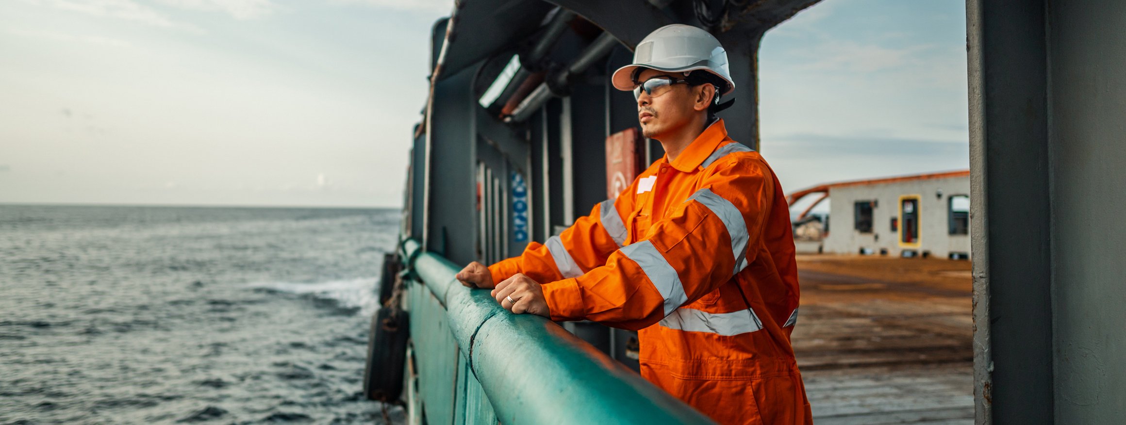 Ein Arbeiter mit Helm und Arbeitskleidung steht an der Reling auf einem Schiff und schaut auf das Meer