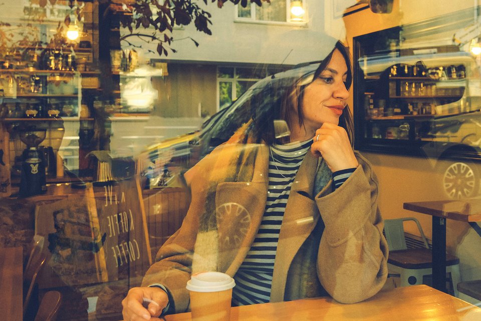 Eine junge Frau sitzt entspannt am Fenster eines Cafés, vor ihr steht ein Coffee-to-go