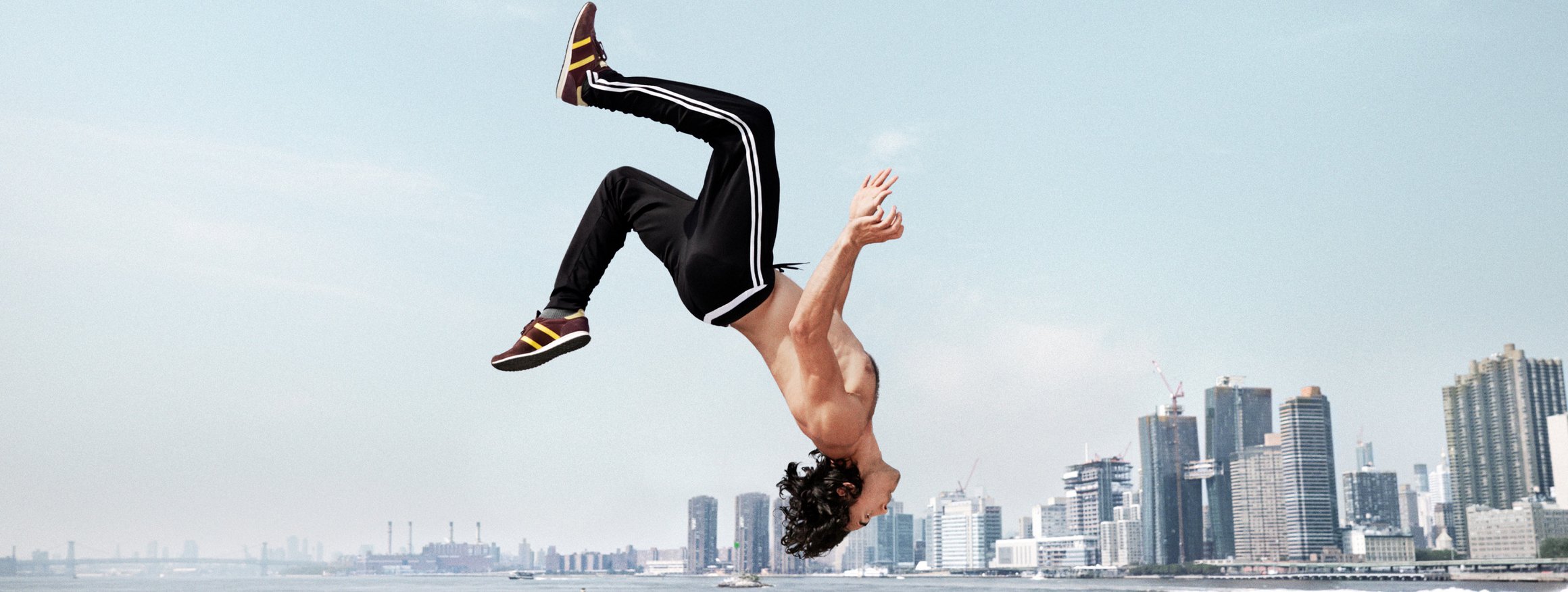 Ein Mann mit Sporthosen, Sportschuhen und nacktem Oberkörper macht einen riskanten Salto an einem Fluss, dahinter die Skyline einer großen Stadt
