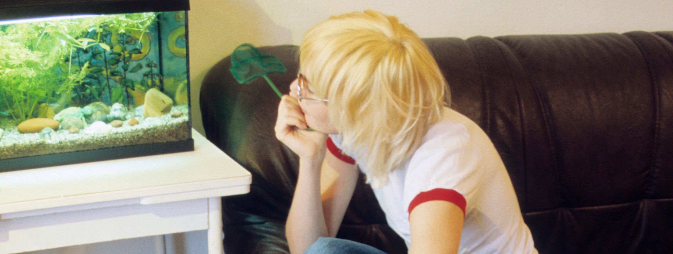 Eine junge blonde Frau im T-Shirt sitzt auf einem Sofa und schaut auf verträumt auf ein Aquarium, das neben ihr auf einem Tisch steht