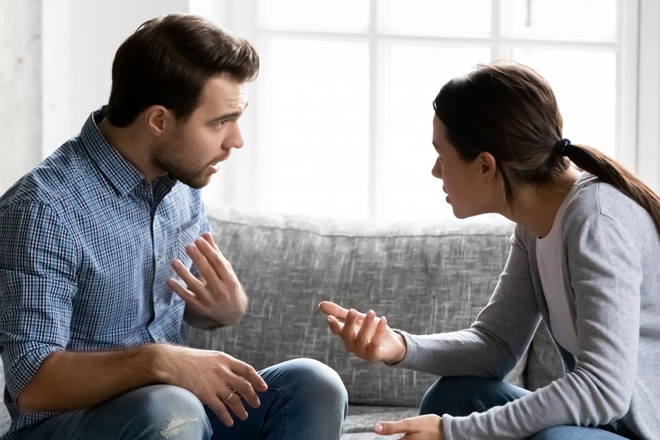 Junges Paar sitzt diskutierend auf dem Sofa und der Mann bittet um Verständnis