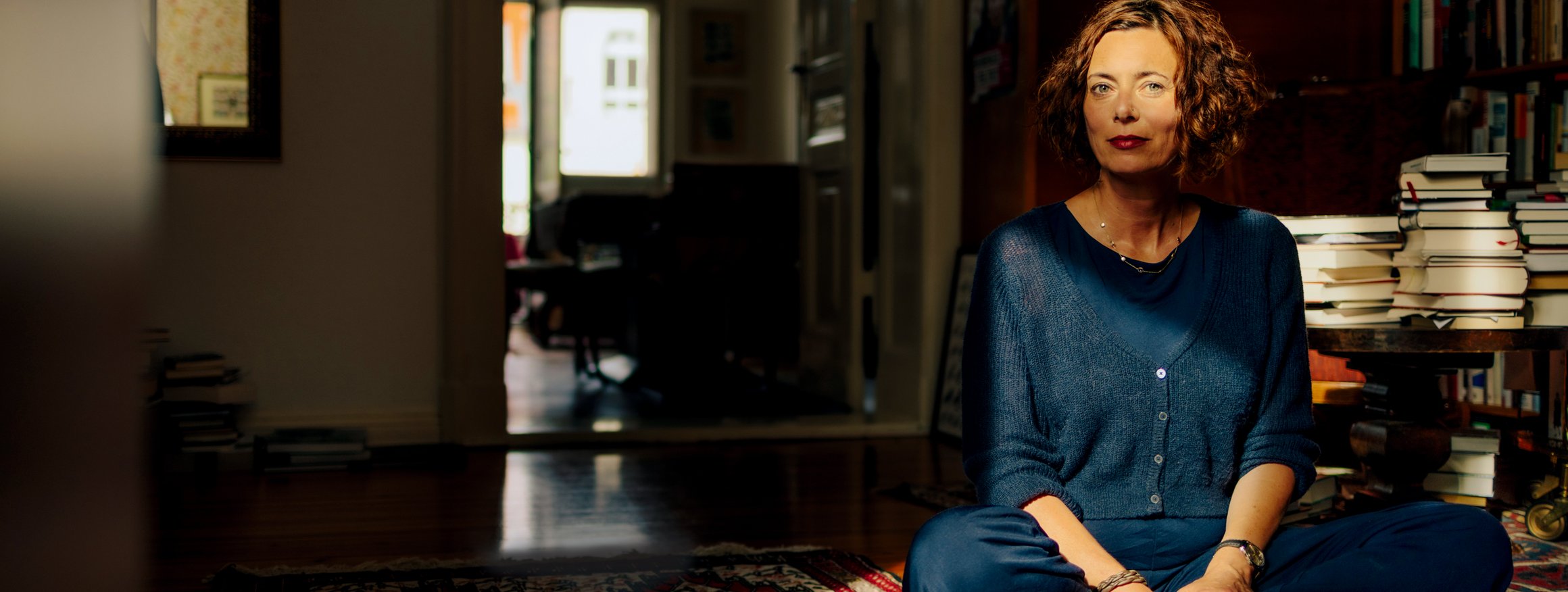 Die Schriftstellerin Eva Menasse sitzt im Schneidersitz auf dem Boden in einer Altbauwohnung, hinter ihr mehrere Stapel Bücher