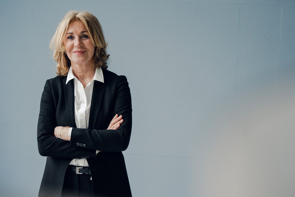 Eine blonde ältere Frau steht in einem dunklem Hosenanzug und verschränkten Armen an einer Wand und lächelt dabei