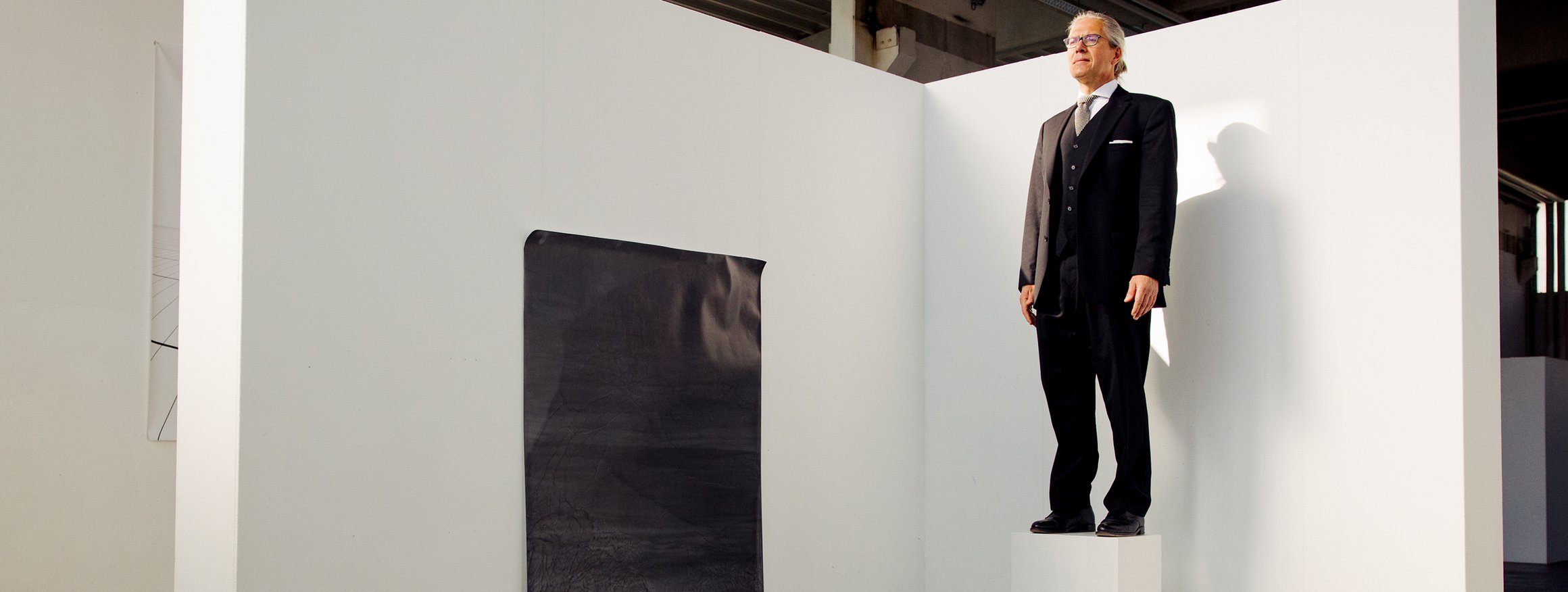 Der Schriftsteller, Steffen Kopetzky, steht in einer Kunstgalerie auf einem Podest wie eine Skulptur, daneben hängt ein Kunstwerk an der Wand