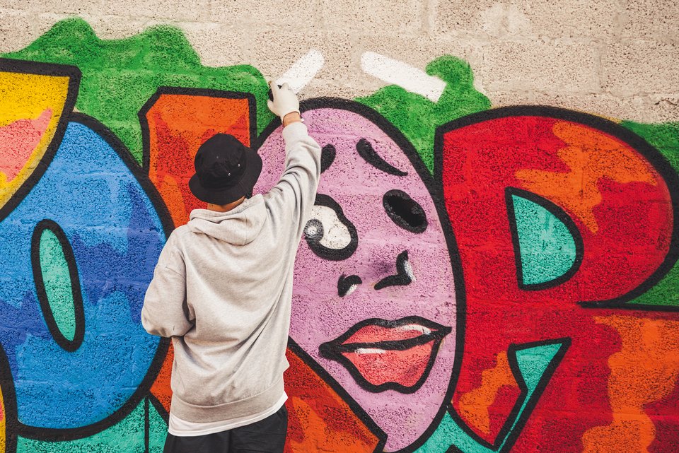 Ein Graffiti-Sprayer mit Mütze und Handschuhen steht an einer Wand und malt mit der Sprühdose ein Bild