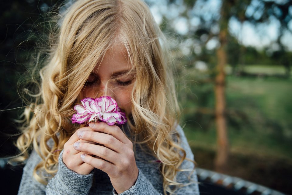 Ein blondes Mädchen mit langen Locken riecht an einer Blume und hat dabei die Augen geschlossen