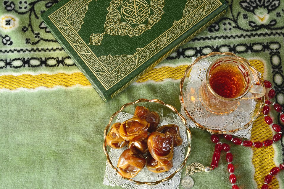 Auf einem grünen orientalischen Teppich liegen der Koran, ein Glas Tee und eine Glasschale mit Datteln