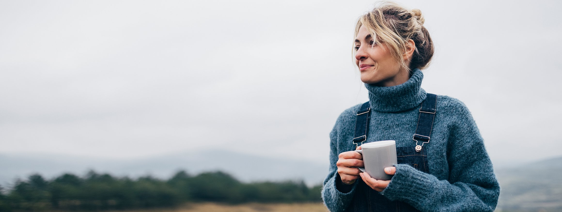 Eine blonde Frau mit hochgesteckten Haaren trägt einen Pullover und eine Latzhose. In der Hand hält sie eine Tasse Tee und lächelt sanft.